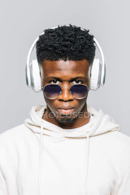 Happy young African American hipster guy in white hoodie and trendy sunglasses listening to music through wireless headphones — Stock Photo