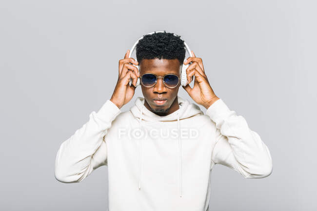 Glücklicher junger afroamerikanischer Hipster in weißem Kapuzenpulli und trendiger Sonnenbrille, der über drahtlose Kopfhörer Musik hört — Stockfoto