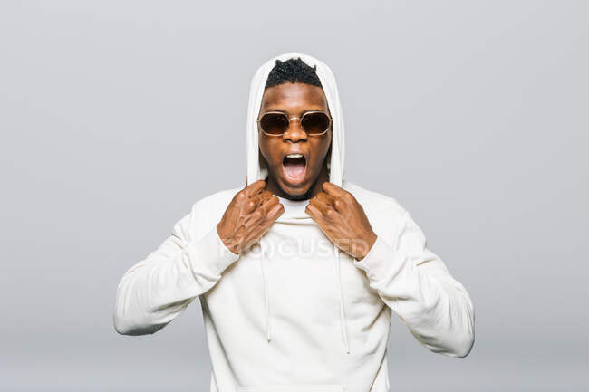 Expressive African American male in white hoodie and stylish sunglasses screaming on gray background in studio — Stock Photo