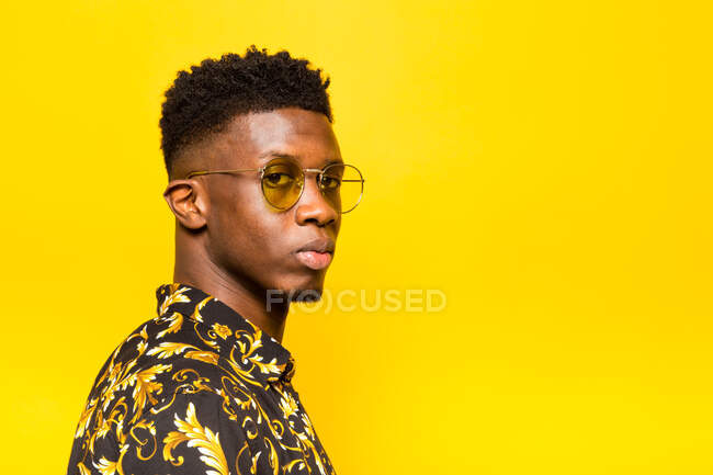 Portrait of unemotional African American male in trendy outfit standing on yellow background in studio looking at camera — Stock Photo