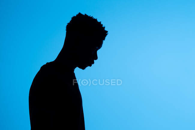 Vista laterale di silhouette di maschio afroamericano irriconoscibile in piedi su sfondo blu in studio scuro — Foto stock