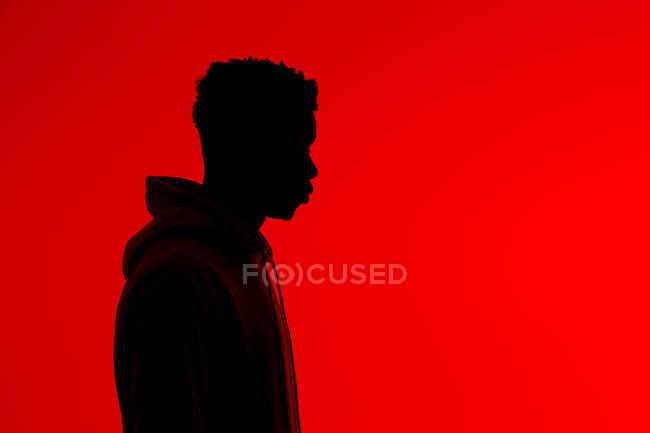 Vista lateral de silueta de varón afroamericano irreconocible con capucha de pie sobre fondo rojo en estudio oscuro - foto de stock