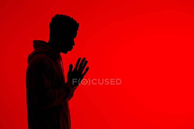 Vista laterale di silhouette di maschio afroamericano irriconoscibile in piedi con le mani strette e pregando su sfondo rosso in studio — Foto stock