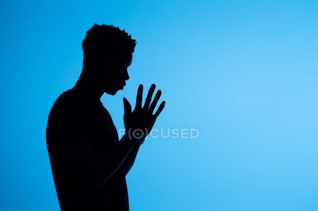 Vista lateral da silhueta de homem afro-americano irreconhecível em pé com as mãos apertadas e orando em fundo azul no estúdio — Fotografia de Stock