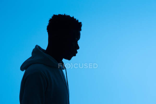 Seitenansicht der Silhouette eines nicht erkennbaren afroamerikanischen Mannes in Kapuzenpulli, der auf blauem Hintergrund im dunklen Studio steht — Stockfoto