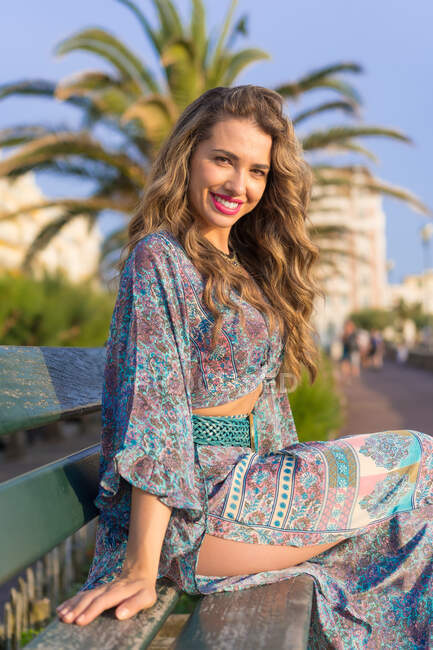 Side view of happy young fashionable female model in colorful boho style  summer outfit sitting on bench in tropical city and looking at camera —  positive, relax - Stock Photo | #527323074