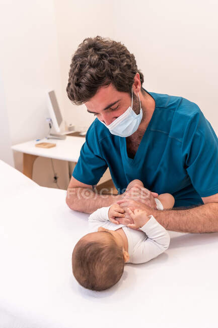 From above of male practitioner in mask examining adorable infant lying on medical table during appointment in hospital — Stock Photo