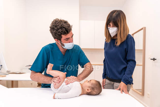 Male pediatrician in mask examining baby in medical room with mother during appointment in modern hospital — Stock Photo