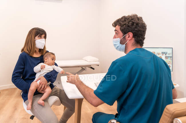 Mère avec bébé mignon assis à la table avec pédiatre expliquant le diagnostic sur ordinateur lors d'une visite dans une clinique moderne — Photo de stock