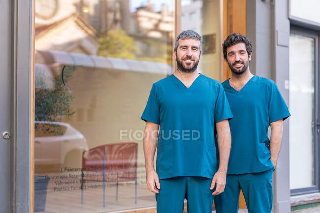 Délicieux praticiens masculins en uniforme bleu debout près de l'entrée de l'hôpital moderne en ville et regardant la caméra — Photo de stock