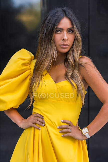 Modelo feminino jovem em vestido amarelo elegante de mãos dadas na cintura e olhando para a câmera no dia de verão na cidade — Fotografia de Stock