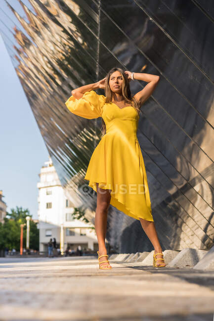 Молода жінка в стильному жовтому вбранні торкається голови і дивиться вгору, стоячи на тротуарі за межами сучасної будівлі в літній день — стокове фото