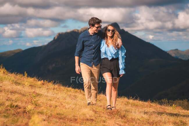 Веселый молодой человек обнимает очаровательную девушку в солнечных очках, взаимодействуя и прогуливаясь по золотой траве на фоне гор — стоковое фото