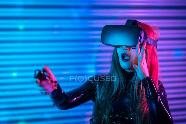 Astonished young woman in VR glasses using controller to interact with virtual reality while playing videogame — Stock Photo