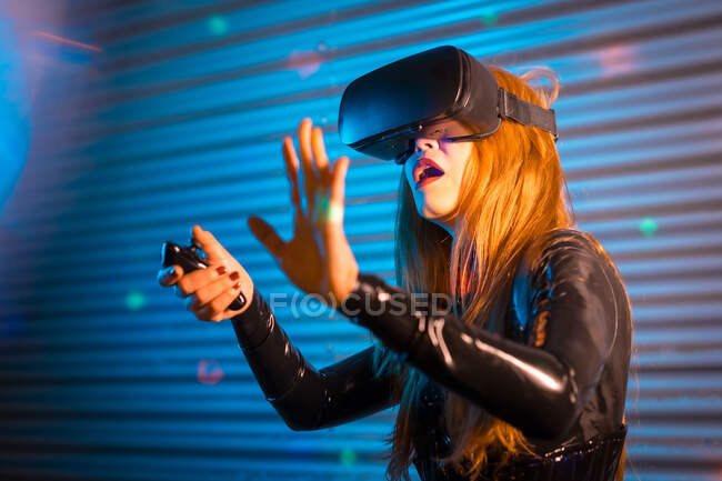 Astonished young woman in VR glasses using controller to interact with virtual reality while playing videogame — Stock Photo
