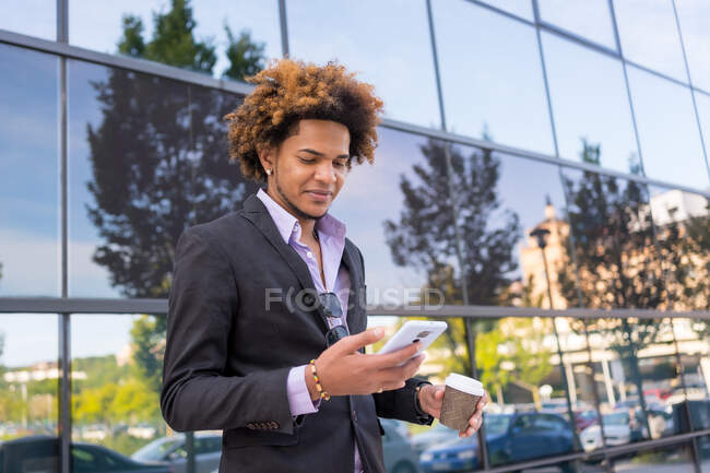 Well dressed African American male entrepreneur browsing mobile phone while walking with takeaway coffee in city street — Stock Photo