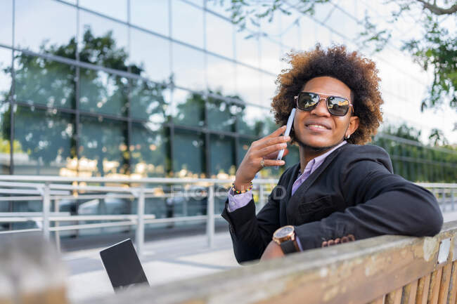 Positive African American male entrepreneur in formal suit and sunglasses with Afro hairstyle speaking on mobile phone while working in city street — Stock Photo