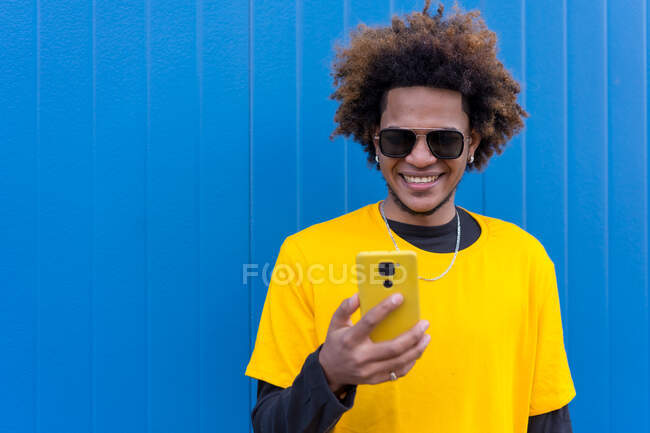 Content African American male with Afro hair talking on mobile phone while standing with takeaway coffee against blue background — Stock Photo