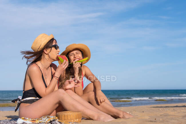 Amici donne spensierate in costume da bagno seduti sulla spiaggia di sabbia e mangiare succosa anguria mentre si godono le vacanze estive — Foto stock