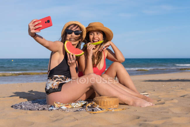 Felice amiche in costume da bagno seduti sulla spiaggia sabbiosa e mangiare anguria dolce mentre scattano autoritratto su smartphone durante le vacanze estive — Foto stock