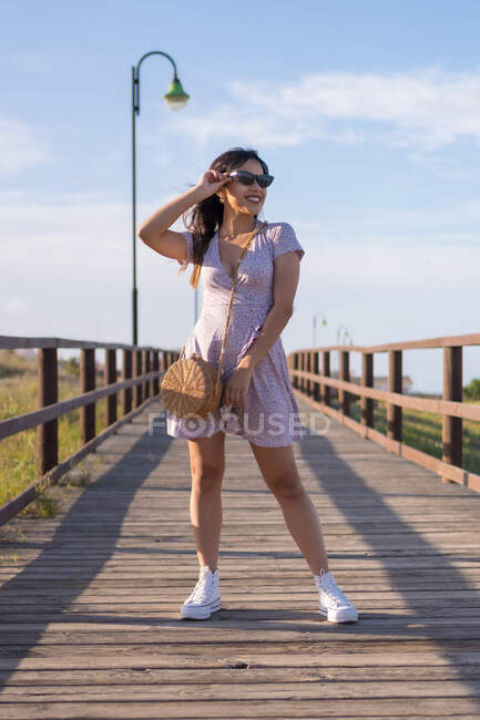 Mulher alegre em vestido e óculos de sol em pé no calçadão e olhando para longe durante as férias de verão — Fotografia de Stock