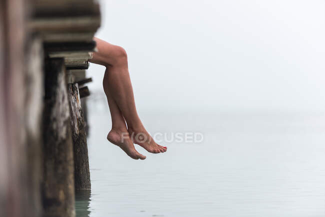 Вид сбоку на ноги женщины, сидящей на деревянной пристани у моря в туманное утро на площади Плайя-де-Муро — стоковое фото