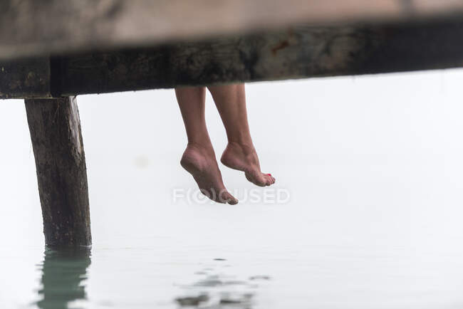 Crop legs of female sitting on wooden quay near sea on misty morning on Playa de Muro — Stock Photo