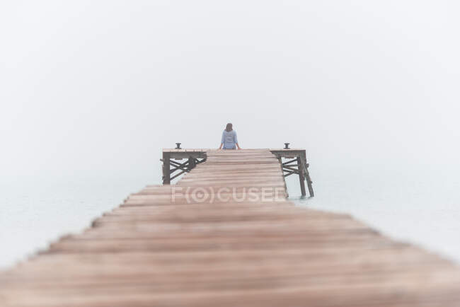 Вид сзади на неузнаваемую женщину, сидящую на деревянном пиале у моря туманным летним утром на площади Playa de Muro в Испании — стоковое фото