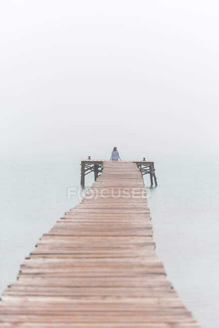 Rückansicht einer unkenntlichen Frau, die an einem nebligen Sommermorgen an der Playa de Muro in Spanien auf einem Holzsteg am Meer sitzt — Stockfoto