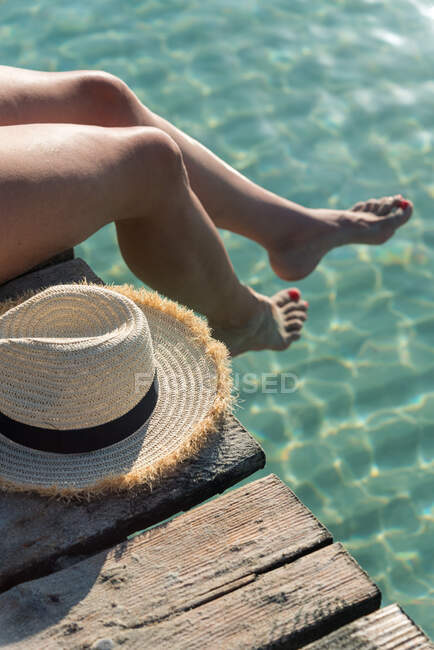 De cima de pernas de colheita de fêmea sentado no cais de madeira com chapéu de palha e desfrutando de férias de verão perto do mar no dia ensolarado na Playa de Muro — Fotografia de Stock