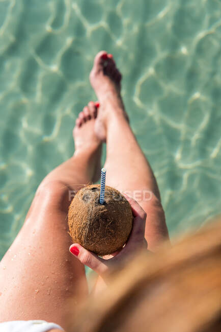 Сверху неузнаваемая женщина сидит с кокосовым коктейлем с соломой возле синего волнистого моря и наслаждается летним отдыхом на Плайя-де-Муро — стоковое фото