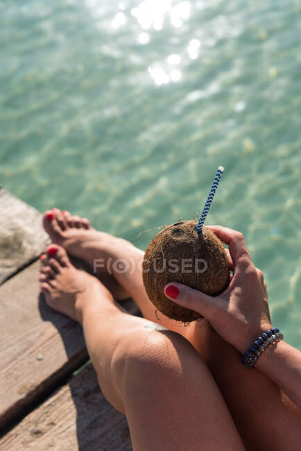 Сверху неузнаваемая женщина сидит с кокосовым коктейлем с соломой возле синего волнистого моря и наслаждается летним отдыхом на Плайя-де-Муро — стоковое фото