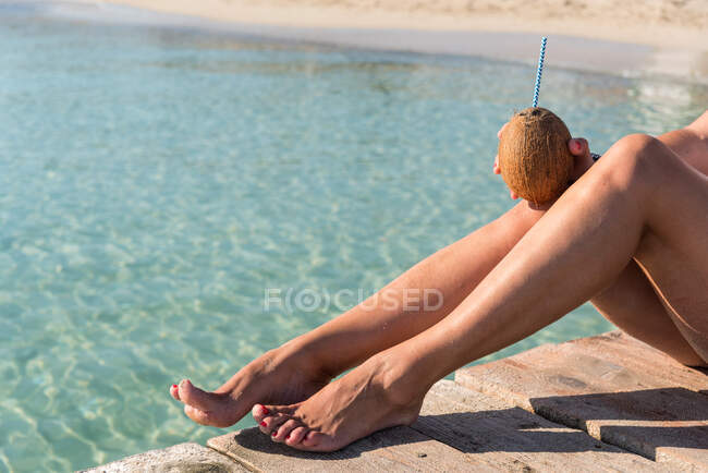 Неузнаваемая женщина сидит с кокосовым коктейлем с соломой возле синего волнистого моря и наслаждается летним отдыхом на Плайя-де-Муро — стоковое фото