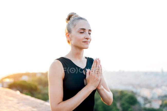 Спокойная женщина с руками на груди и закрытыми глазами медитирует на крыше во время занятий йогой вечером — стоковое фото