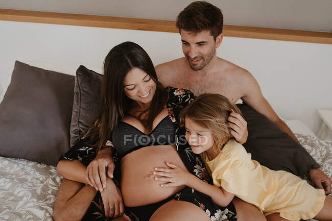 Дитина погладжує животик усміхненої майбутньої мами в нижній білизні, лежачи на ліжку з чоловіком в будинку — стокове фото