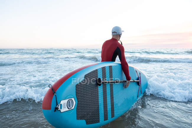 Vista posterior de surfista masculino irreconocible en traje de neopreno y sombrero llevando tabla de paddle y entrando al agua para surfear en la orilla del mar - foto de stock