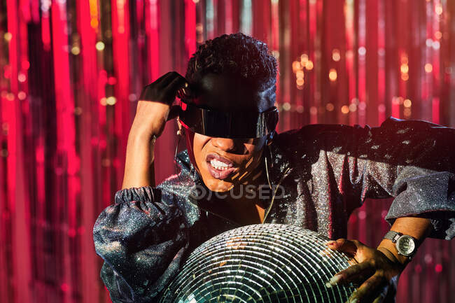 Модная этническая женщина в киберпанк-очках и наручных часах, улыбающаяся при прикосновении к диско-мячу в световом луче — стоковое фото