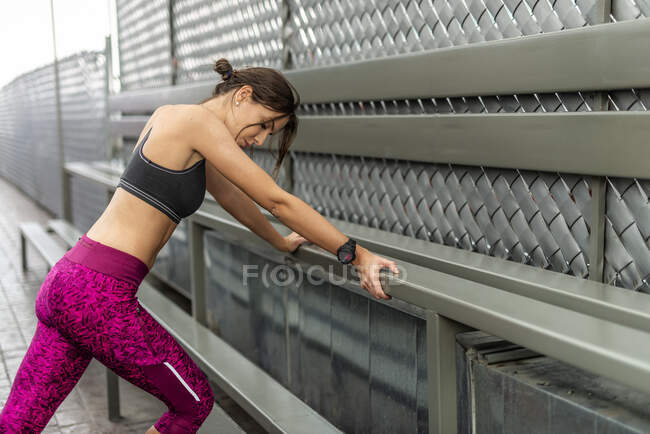Vista lateral de la atleta cansada sudada en ropa deportiva apoyada en el banco durante el descanso en el entrenamiento al aire libre - foto de stock