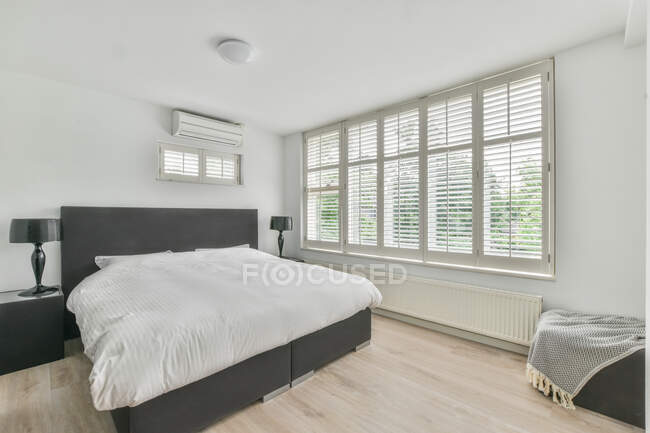 Удобная мягкая кровать с белым одеялом, расположенная рядом с большими окнами в современной спальне в квартире — стоковое фото