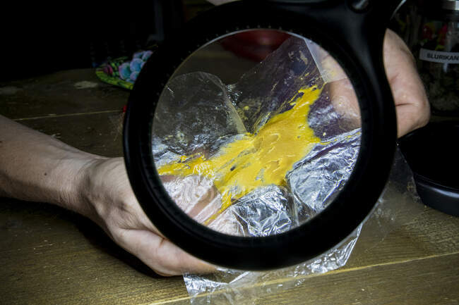 Анонимный самец, демонстрирующий марихуанный клей на пластиковом куске через лупу в комнате — стоковое фото
