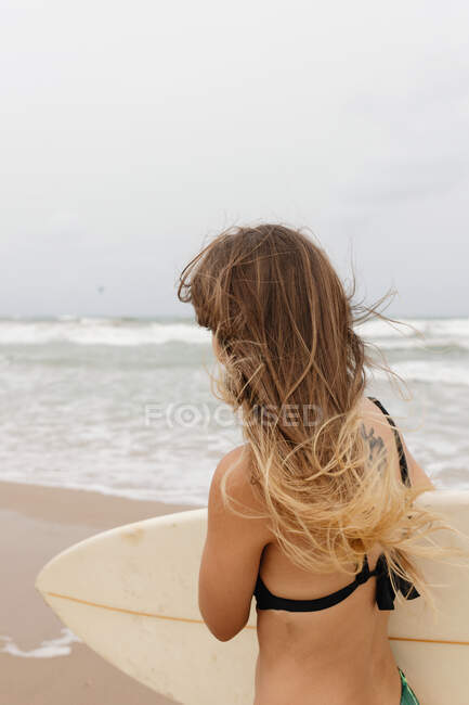 Side view of unrecognizable young sportswoman in swimwear with surfboard looking away on sandy coast against stormy ocean — Stock Photo