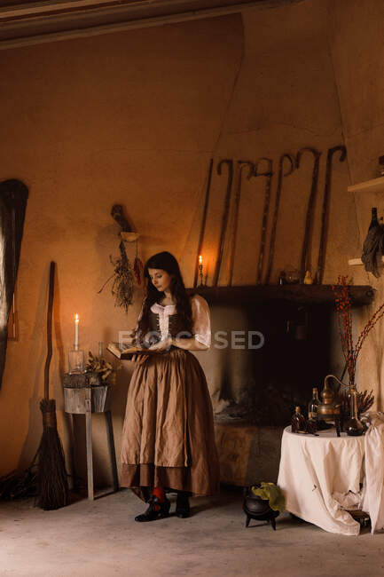Witch in long dress reading magic book of spells while standing in cozy room with broom and cauldron — Stock Photo