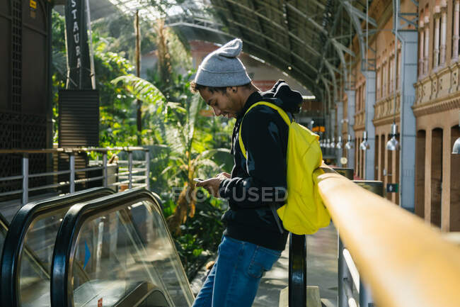 Vue latérale de ravi afro-américain hipster mâle avec sac à dos debout près de l'escalator dans le centre commercial et le surf téléphone mobile — Photo de stock