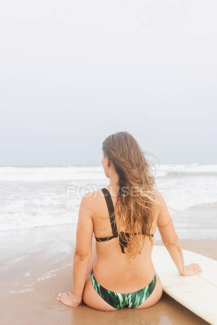 Задний вид анонимной спортсменки в купальнике с доской для серфинга, сидящей на песчаном побережье, созерцающей океан под легким небом — тело, Пена - Stock Photo