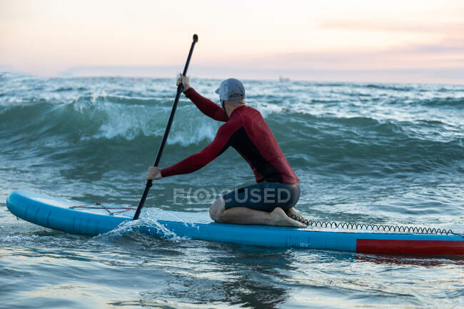 Side view of male surfer in wetsuit and hat on paddle board surfing on seashore — Stock Photo