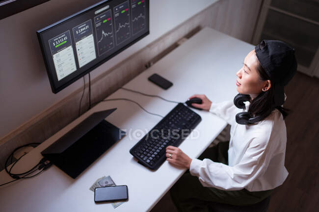 Високий кут зосереджує азіатську жінку, яка працює на комп'ютері з графіками, що показують динаміку змін в вартості криптовалют на зручному робочому місці. — стокове фото