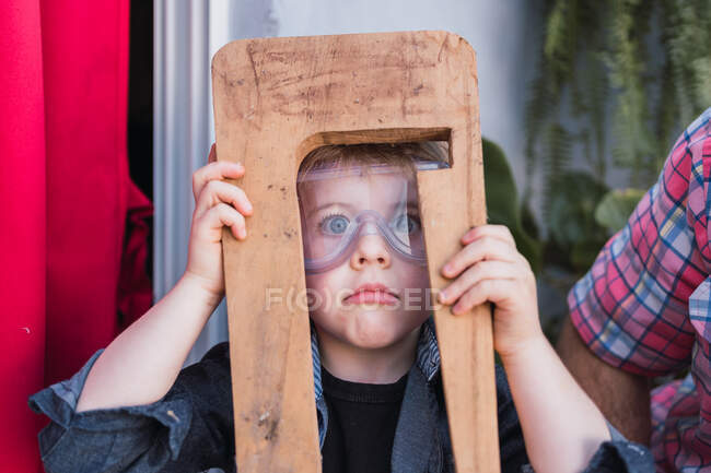 Enfant étonné dans des lunettes de sécurité avec pièce en bois regardant la caméra contre la récolte papa méconnaissable en plein jour — Photo de stock