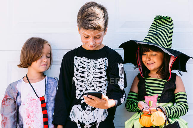 Corps complet de groupe de petits enfants vêtus de divers costumes d'Halloween avec Jack O Lantern sculpté navigation téléphone mobile ensemble tout en se tenant près du mur blanc sur la rue — Photo de stock