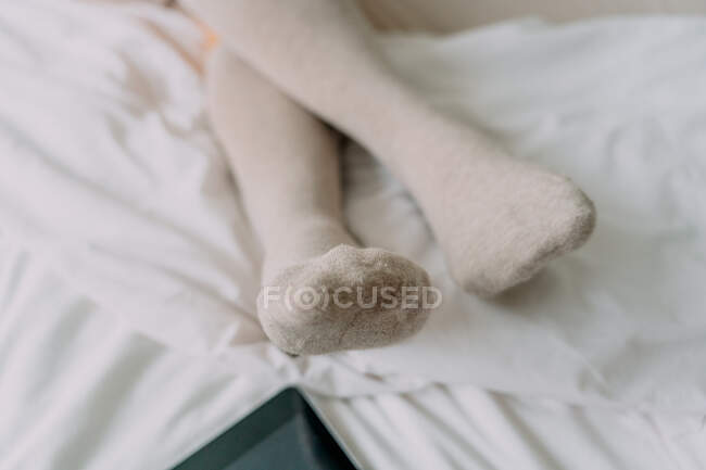 Crop unrecognizable female in knee socks sitting with crossed legs against cellphone with black screen on crumpled bed sheet — Stock Photo