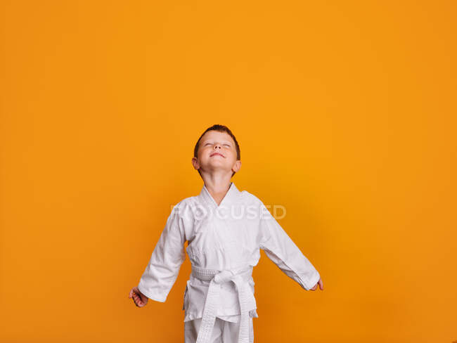 Vista frontal de un niño sonriente en kimono blanco, mirando al techo con los ojos cerrados y los brazos extendidos y las manos en puño. - foto de stock